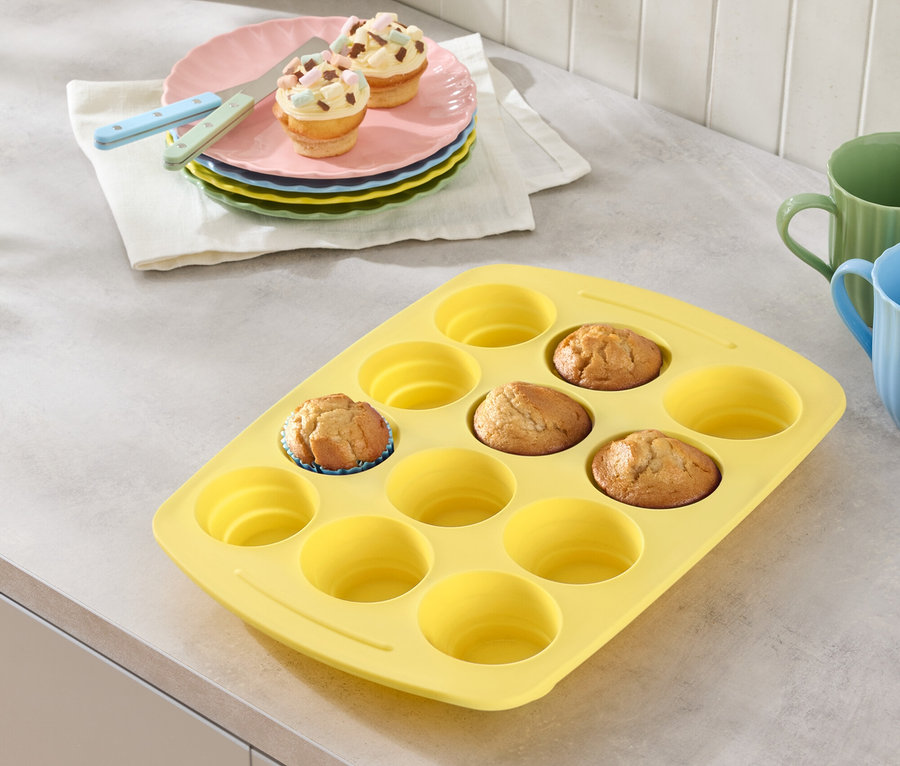 Žltá forma na muffiny s niekoľkými muffiny, taniere s muffiny a poháre na pracovnej doske.