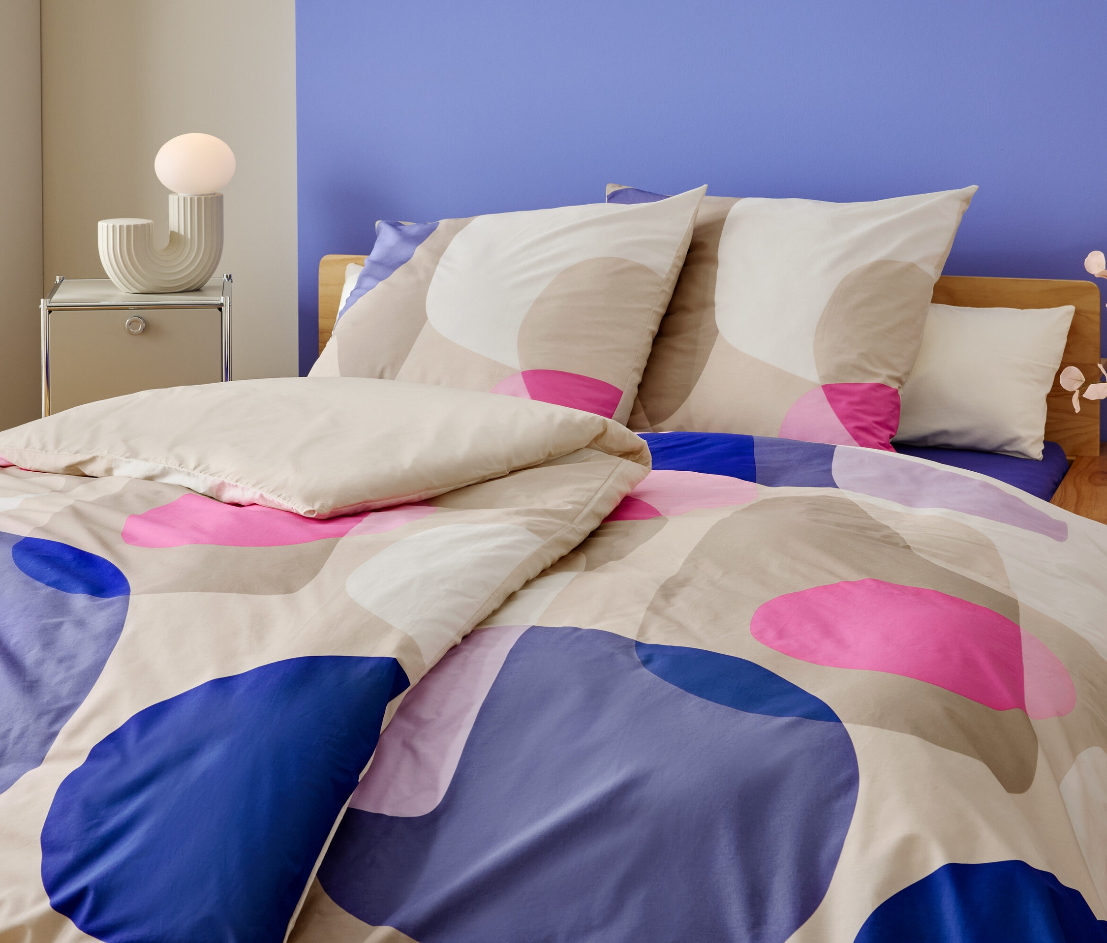 Detailný záber postele s geometrickým vzorom v modrej, ružovej a béžovej farbe. Na nočnom stolíku je lampa a váza s kvetmi.