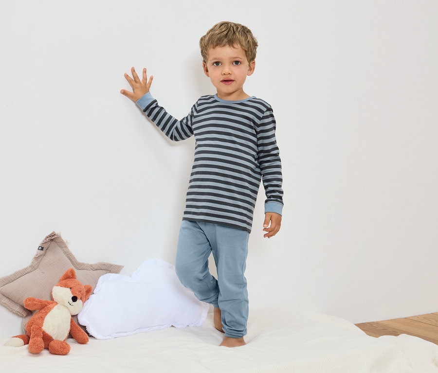 Chlapec stojí v modrom pruhovanom pyžame vedľa plyšovej líšky a vankúšov.