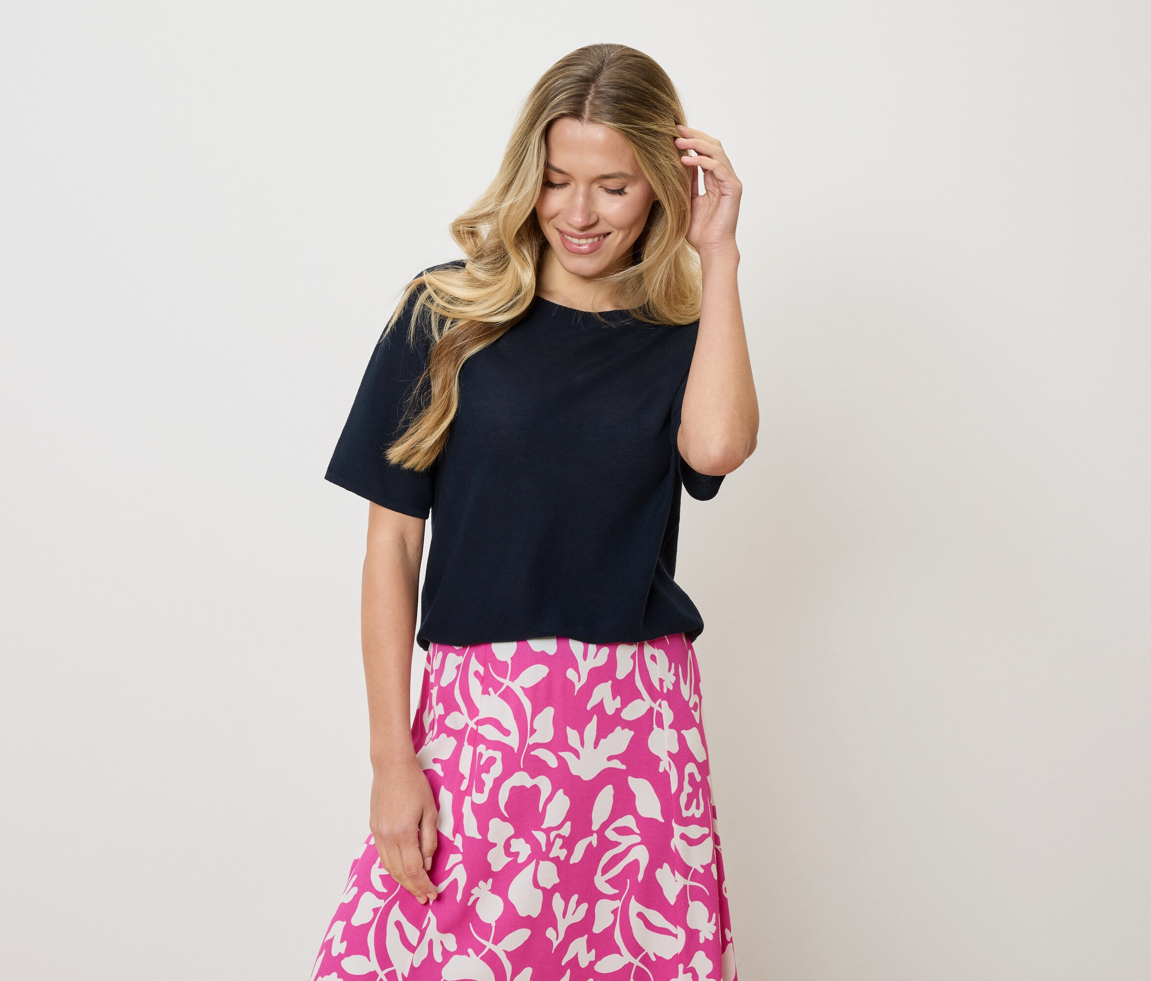 Žena s blond vlnitými vlasmi nosí tmavomodré tričko a ružovo-bielu sukňu s kvetinovou potlačou, pričom sa jednou rukou dotýka vlasov.