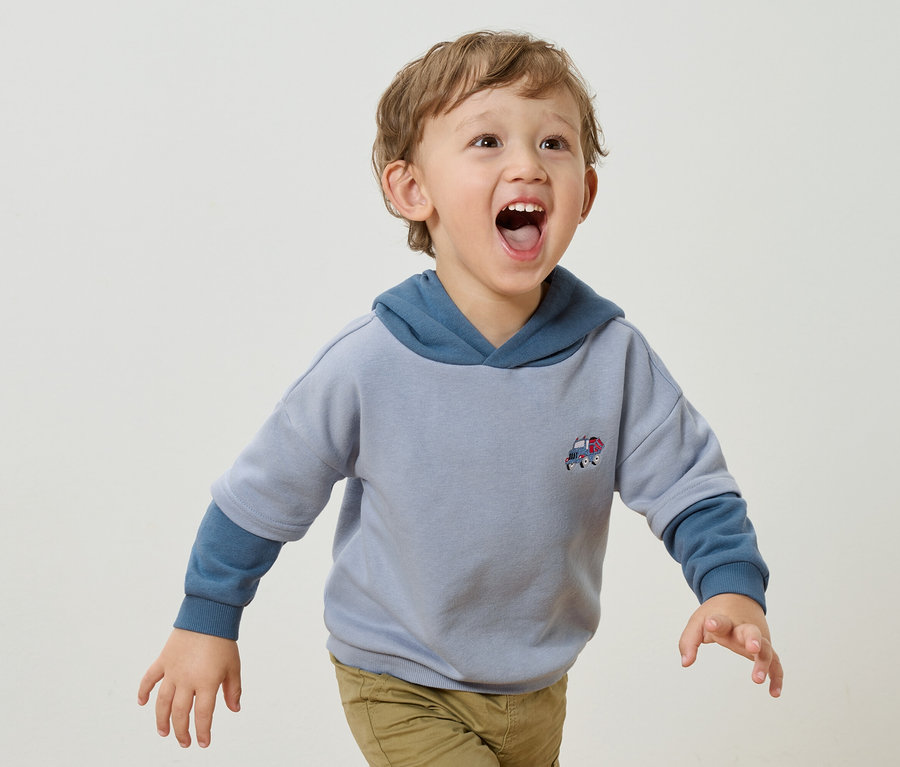 Malý chlapec s blond kučerami a otvorenými ústami má na sebe svetlomodrú mikinu s kapucňou s aplikáciou malého auta a svetlohnedé nohavice na bielom pozadí.