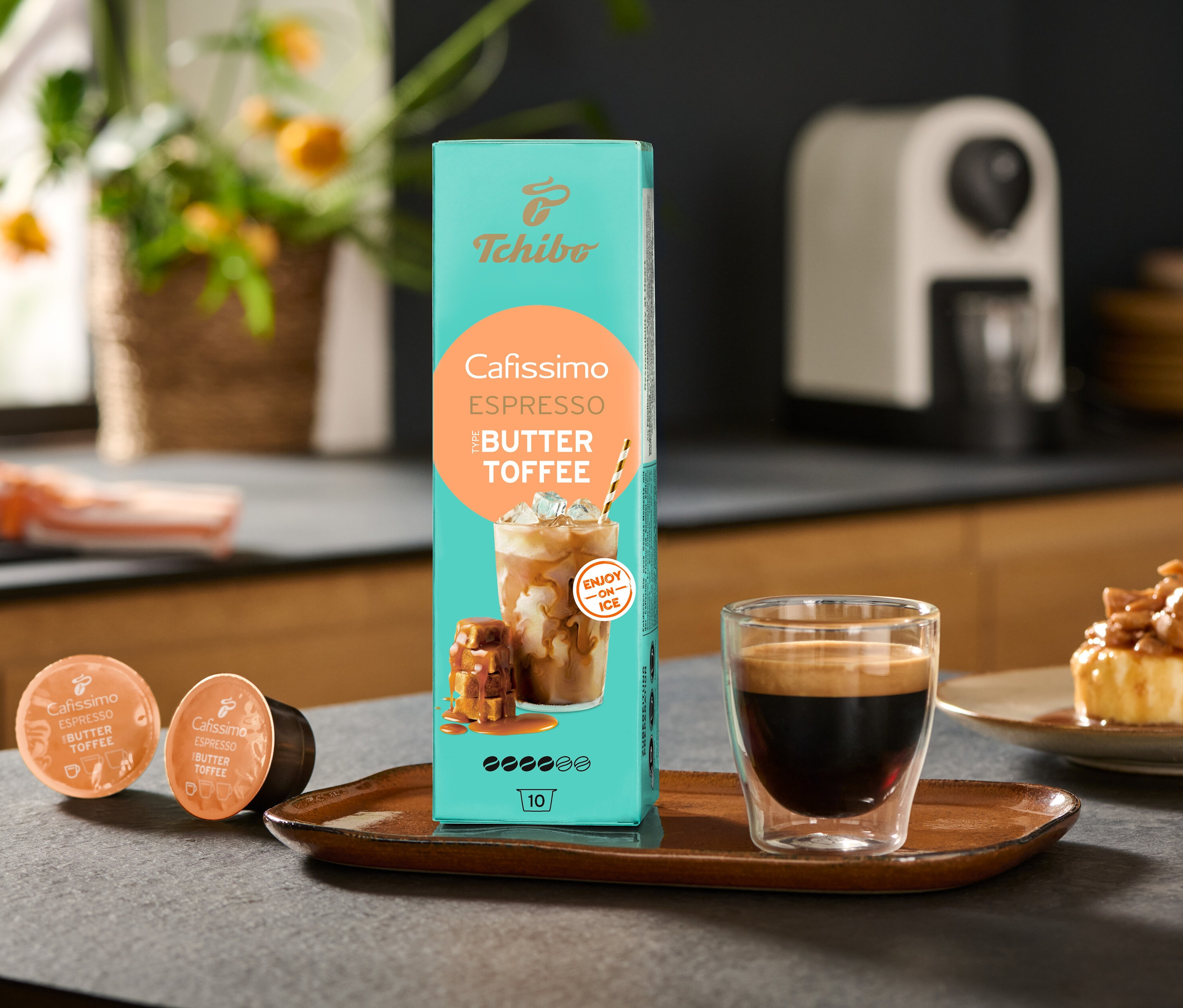Balenie Cafissimo Flavoured Espresso – Buttertoffee a kávovar Cafissimo pure+, white sú na stole spolu so šálkou kávy.