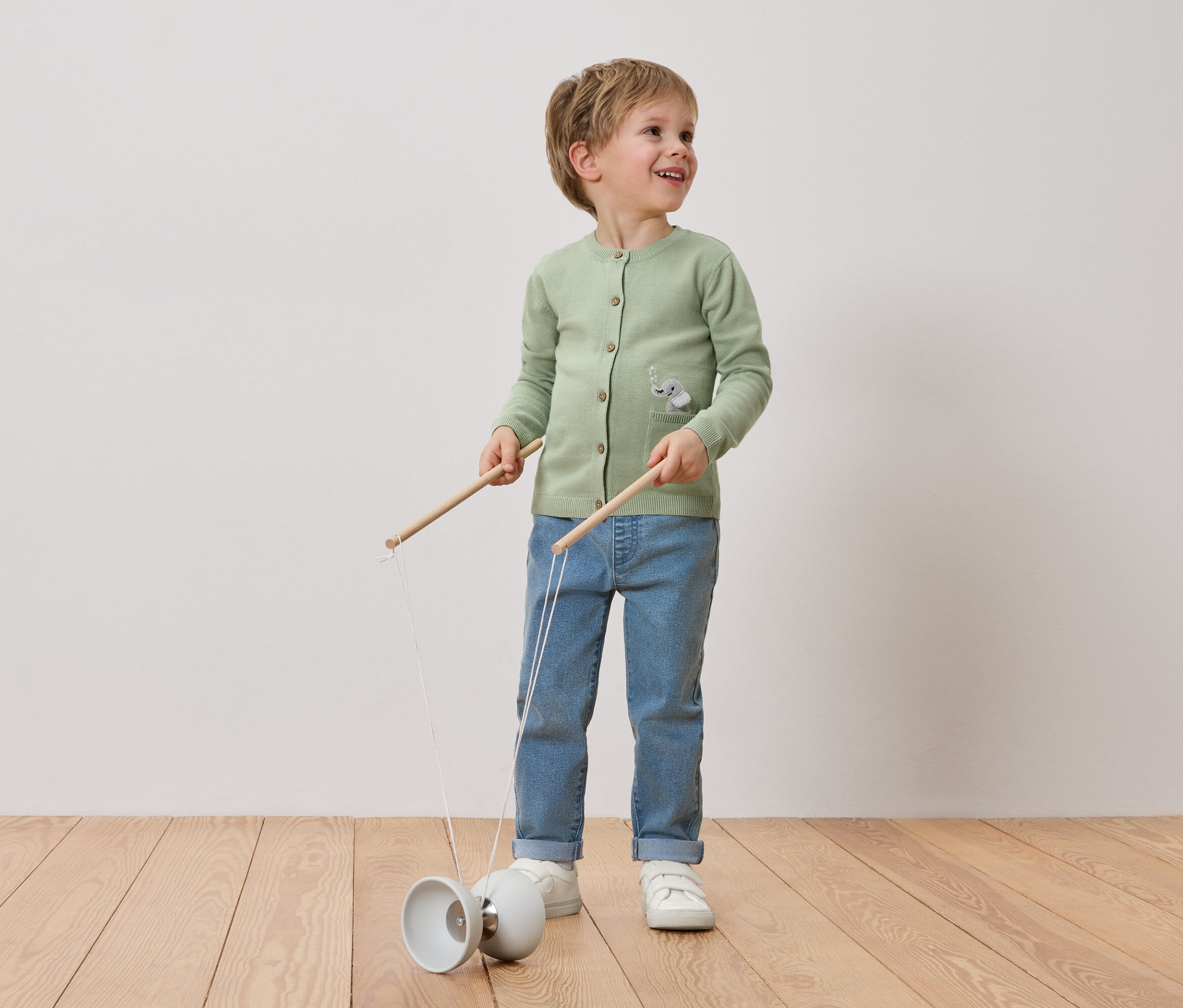 Usmievavý chlapec s blond vlasmi sa hrá s diabolo. Má na sebe zelený kardigán so sloním motívom, džínsy a biele tenisky.