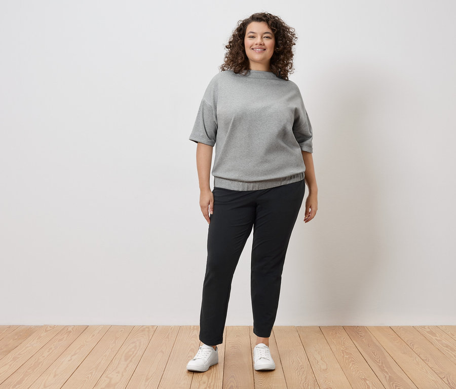 Žena s kučeravými vlasmi stojí pred bielou stenou a má na sebe sivú mikinu, čierne nohavice a biele tenisky.