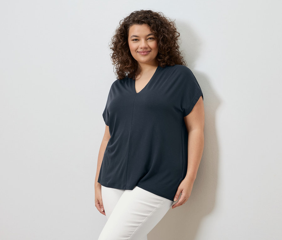 Žena s kučeravými vlasmi stojí pred bielou stenou a má na sebe tmavomodré tričko a biele nohavice.