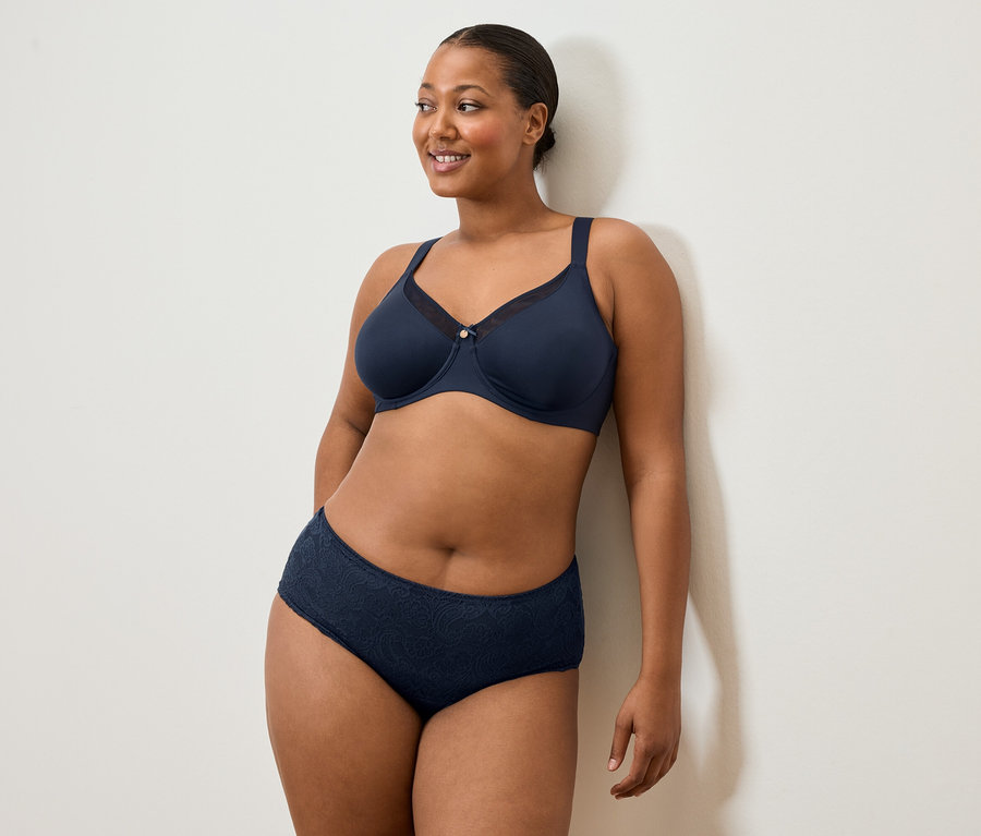 Žena v tmavomodrej podprsenke a nohavičkách stojí pri bielej stene a usmieva sa.