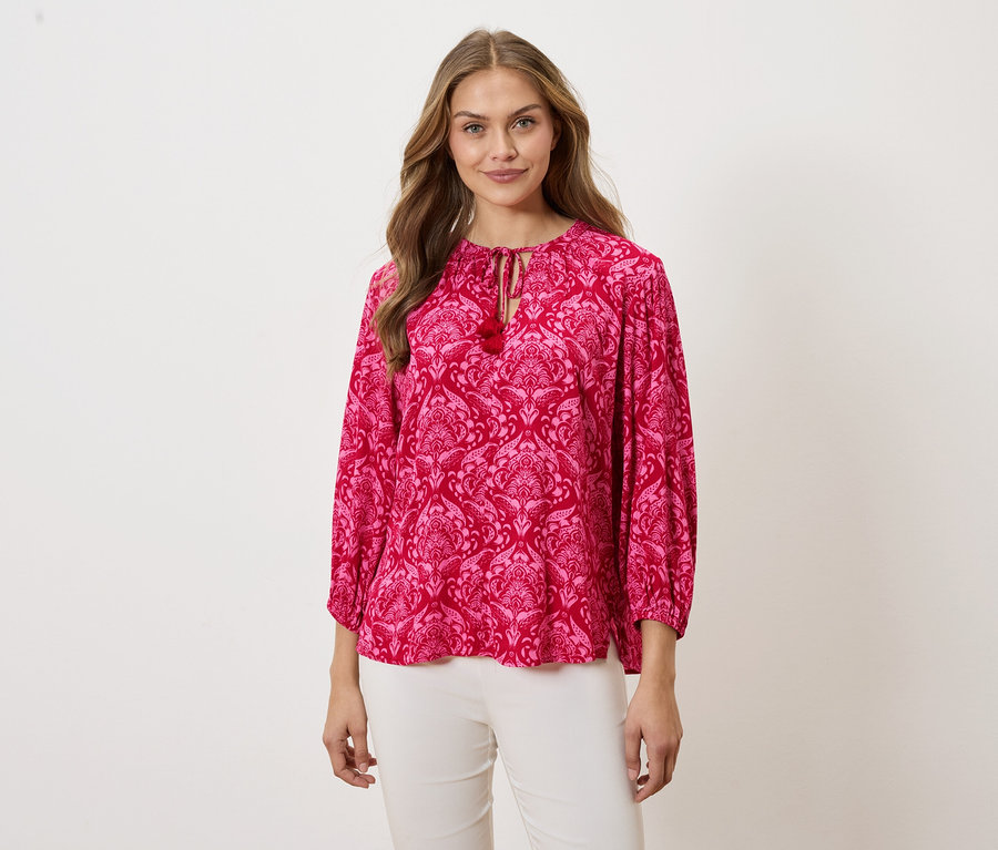 Žena má na sebe ružovú blúzku so vzorom a biele nohavice.