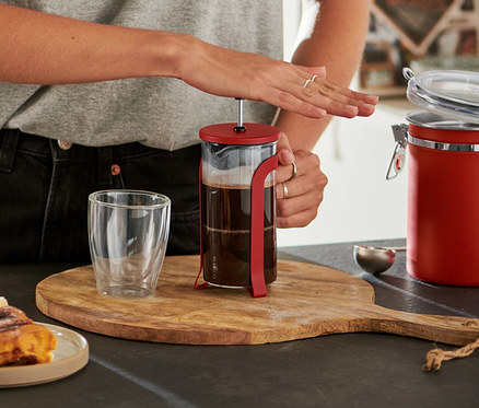 Usmievavá žena v okuliaroch stláča piest červenej kanvice french press 300 ml na drevenej doske, vedľa sú škoricové slimáky a prázdny pohár.