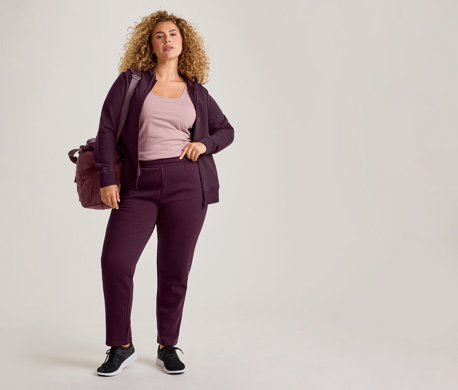 Žena s kučeravými vlasmi má na sebe fialovú mikinu na zips s kapucňou, zladené tepláky, svetloružový top a čierne tenisky, s fialovou športovou taškou cez rameno.
