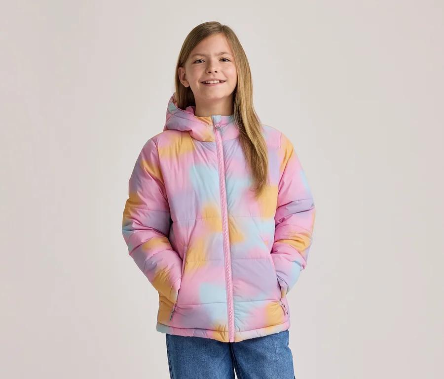 Usmiate dievča v farebnej tie-dye puffer bunde v ružovej, žltej a modrej farbe s kapucňou a ružovým zipsom, ruky vo vreckách.