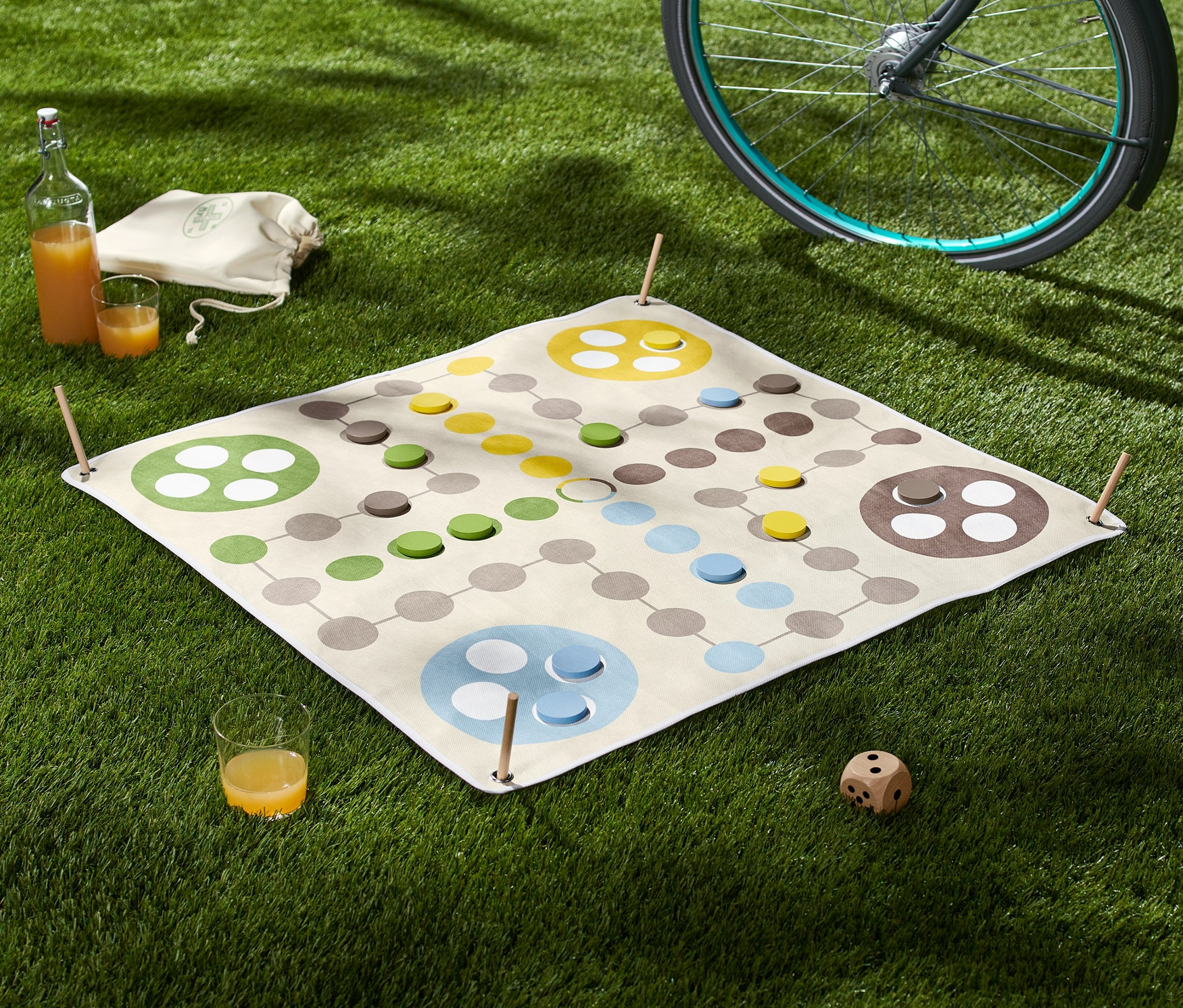 Na tráve leží XL hra Človeče, nehnevaj sa s kockou, vedľa nej je pohár džúsu a fľaša. V pozadí je bicykel.