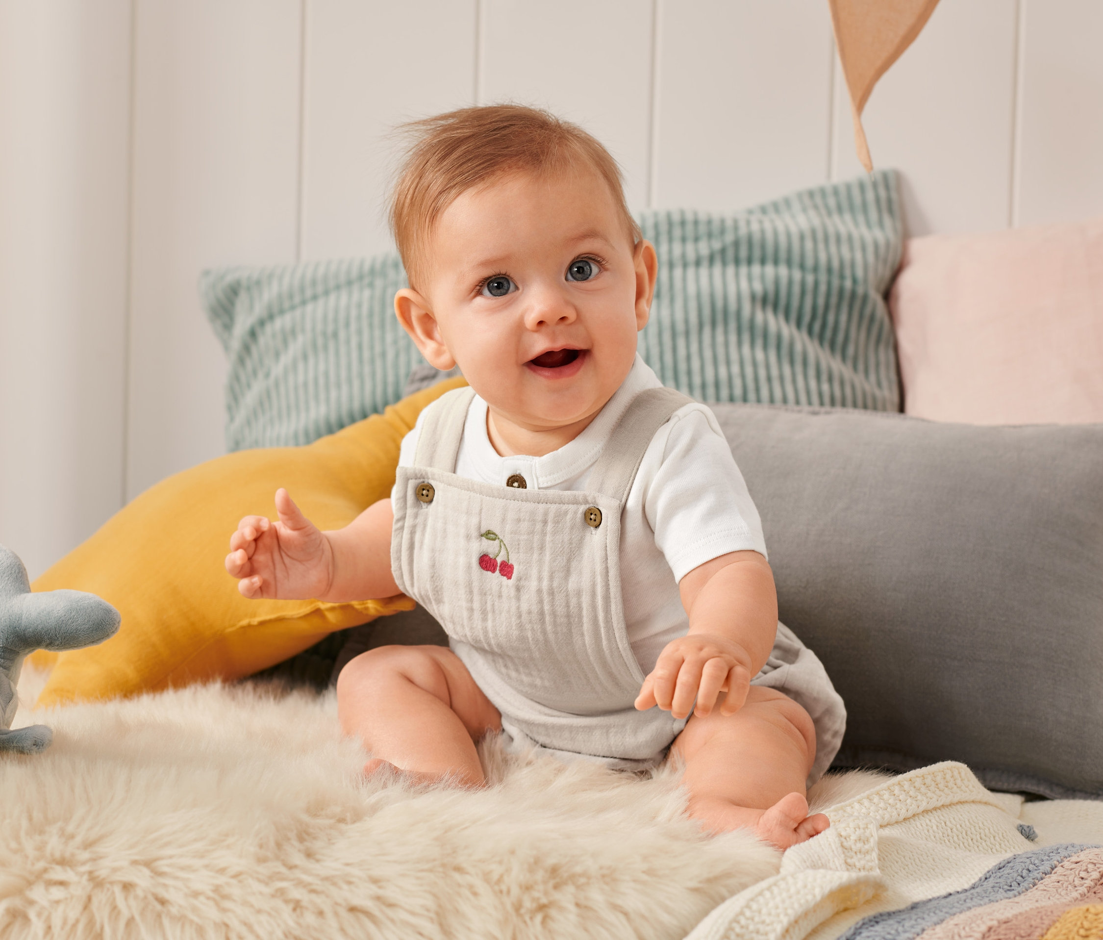 Usmievajúce sa dieťa sedí na kožušine a má na sebe detské body s výšivkou čerešní.