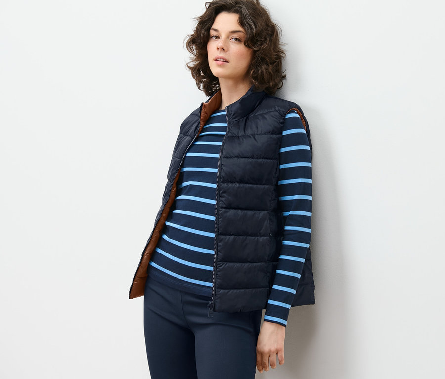 Žena opretá o stenu má na sebe obojstrannú prešívanú vestu, pruhované tričko a nohavice.