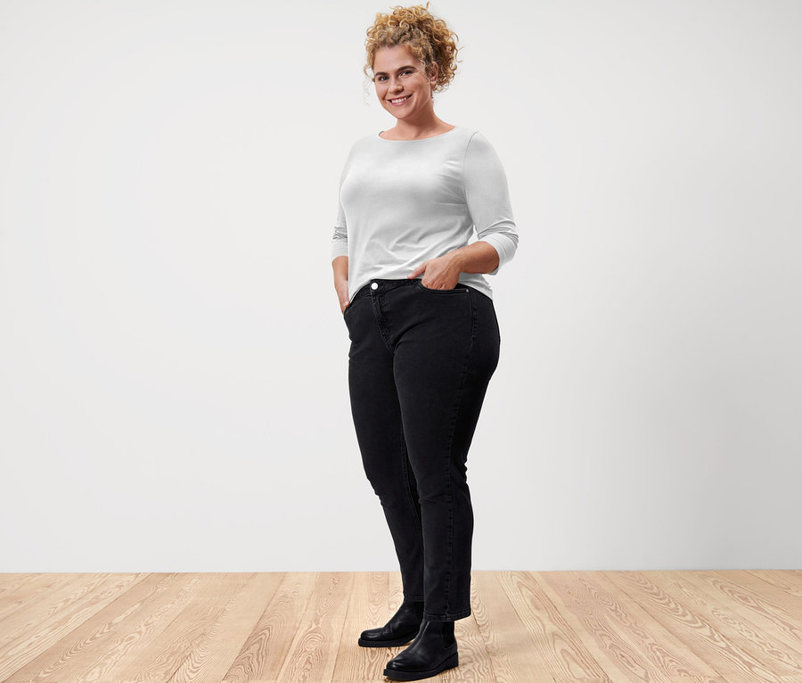 Žena pózuje v bielom tričku s trojštvrťovými rukávmi a čiernych džínsoch.