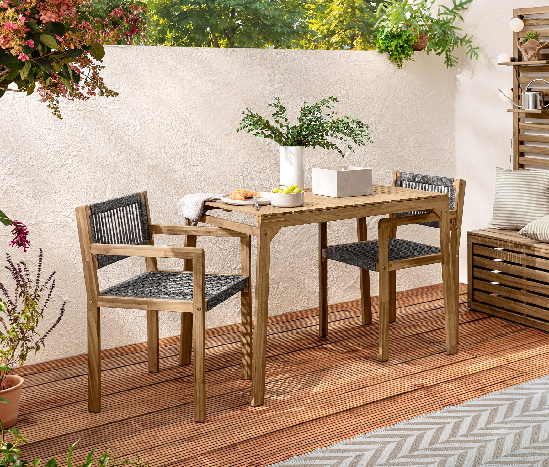 Balkónová súprava s textilným prútím na drevenej terase. Na stole stojí XL LED sviečka z pravého vosku v betónovom kvetináči. Zdobia ho outdoorové vankúše na dotyk pripomínajúce ľan a vonkajšia svetelná reťaz s LED diódami.