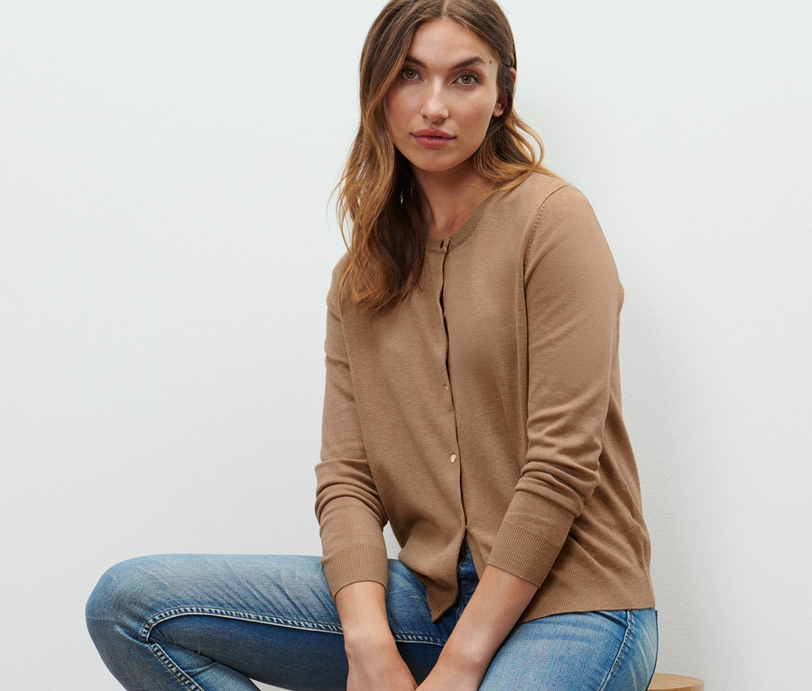 Žena sedí na drevenej stoličke a má na sebe kardigán z jemného úpletu, džínsy a tenisky.