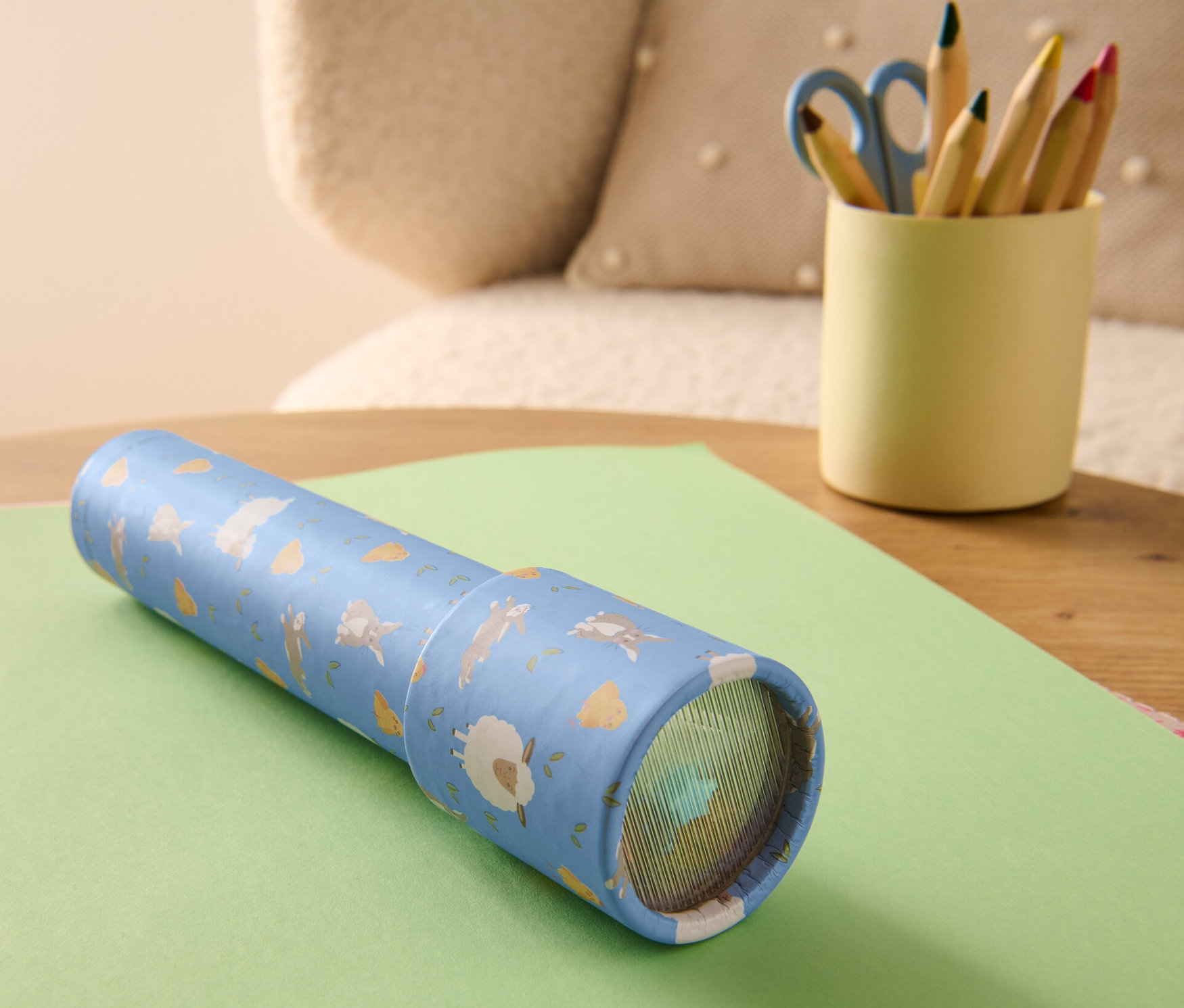 Kaleidoskop leží na zelenom papieri. Vedľa neho stojí téglik s pastelkami a nožnicami.