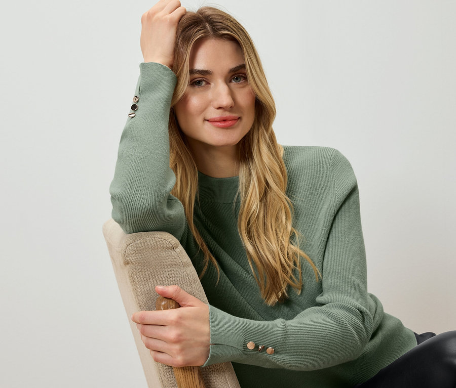 Žena sedí a opiera si hlavu o ruku, má na sebe zelený sveter.