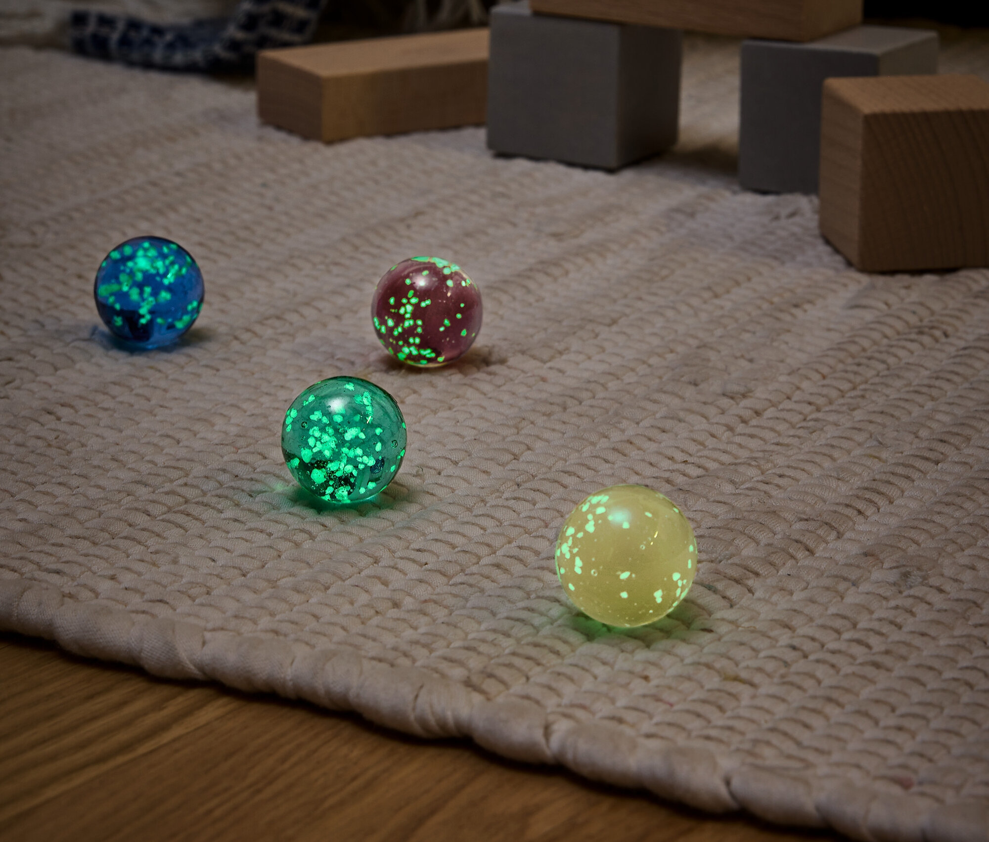 Štyri svietiace gule rôznych farieb (modrá, červená, zelená a žltá) na béžovom koberci s drevenými hračkami v pozadí.