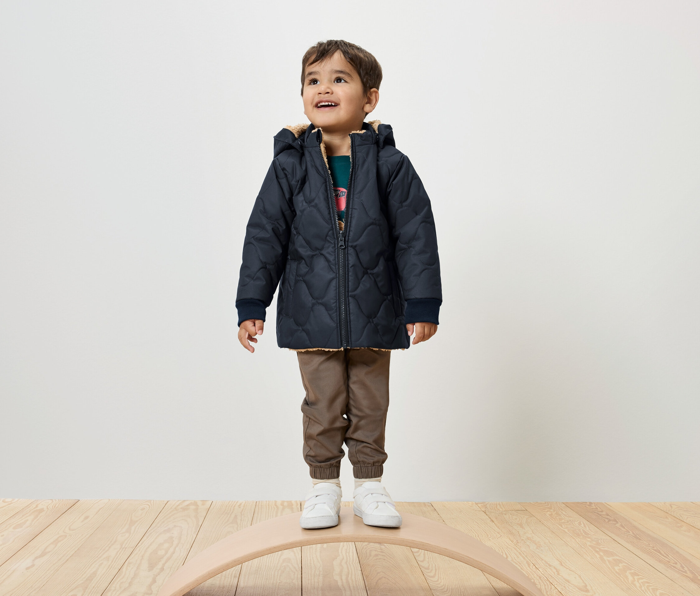 Chlapec stojí na balančnej doske a má na sebe tmavomodrú prešívanú bundu, hnedé nohavice a biele tenisky.