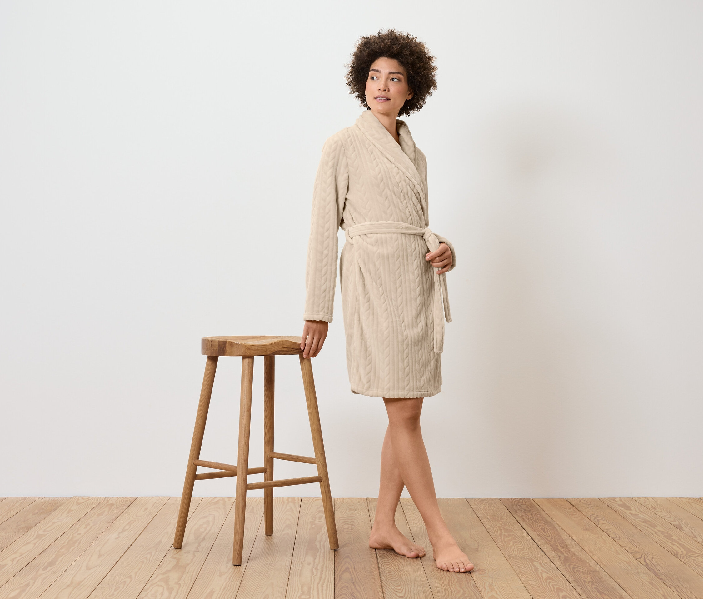 Žena v krémovom župane stojí vedľa drevenej stoličky.