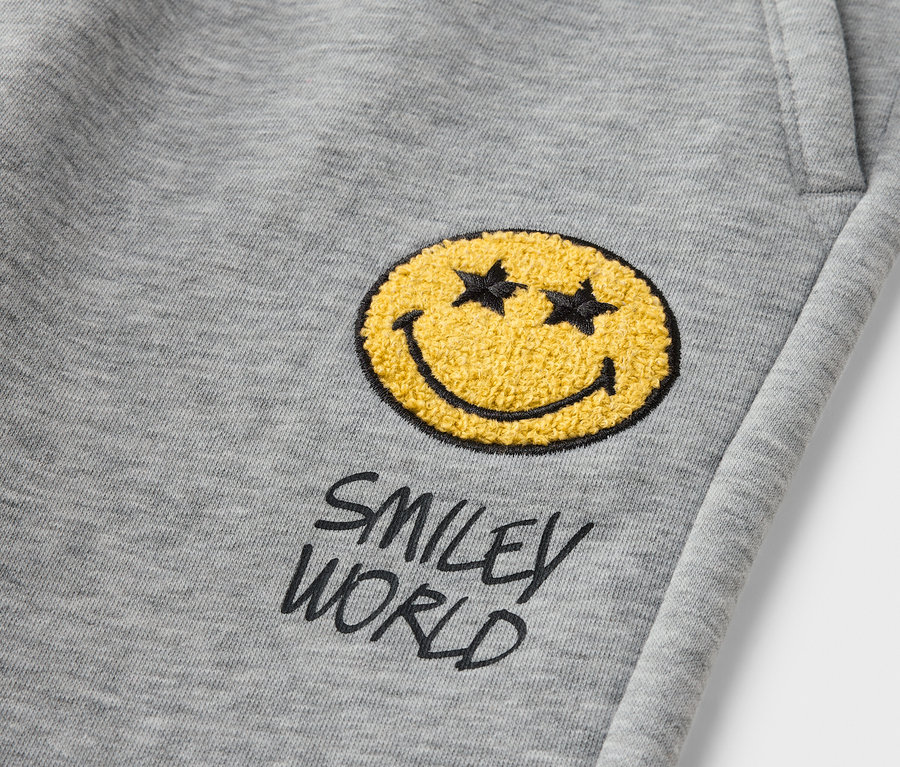 Detail sivých detských joggingových nohavíc SMILEYWORLD® s nášivkou žltého smajlíka.