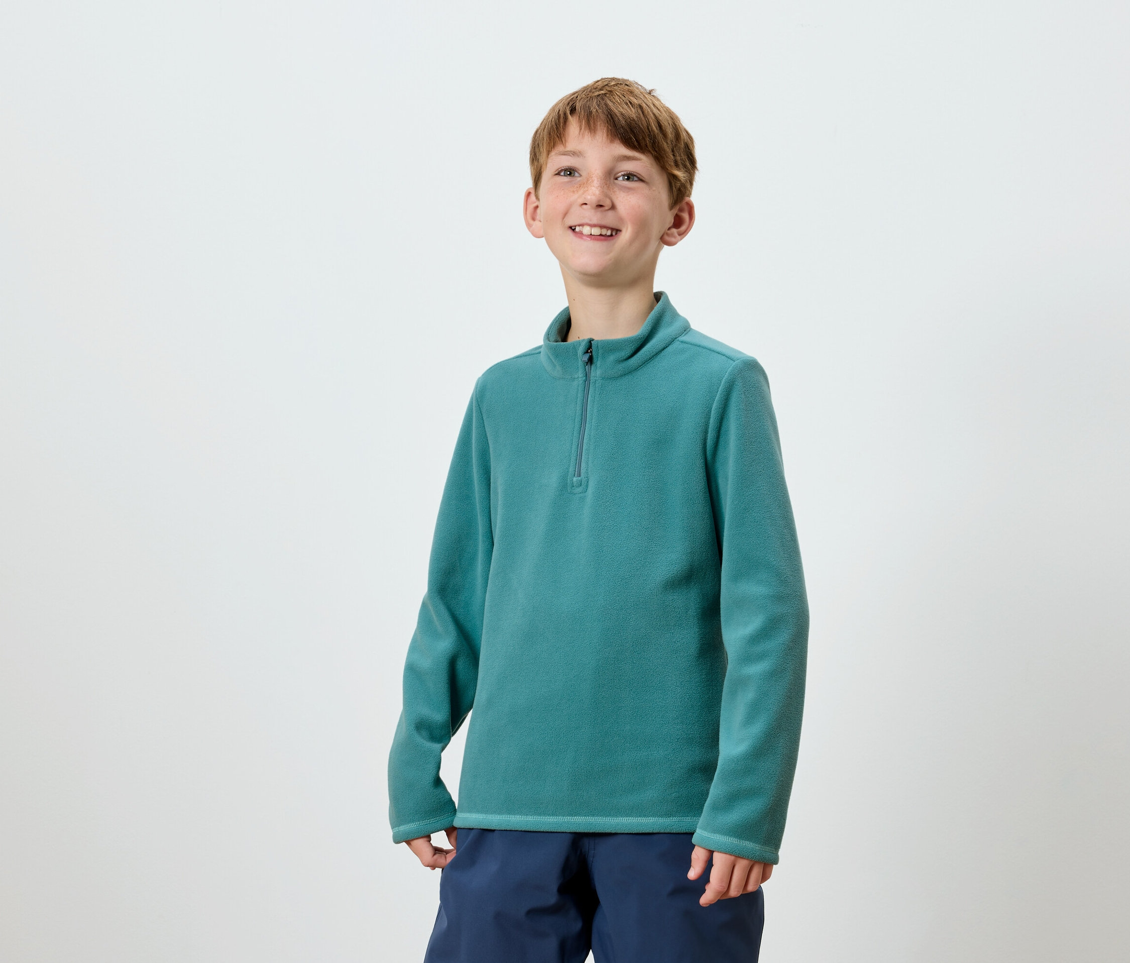 Usmievajúci sa chlapec pózuje v tyrkysovej mikine na zips a tmavomodrých nohaviciach.
