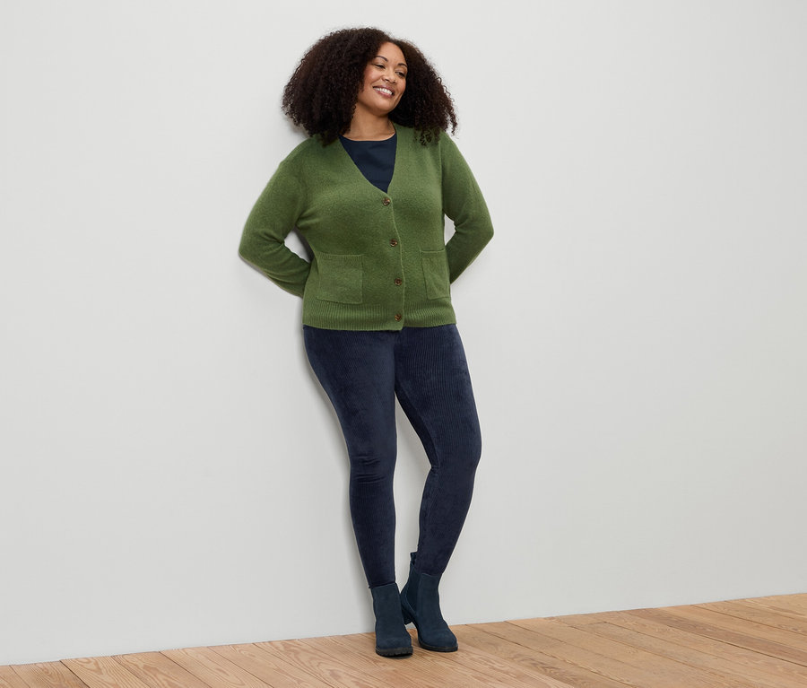 Žena s kučeravými vlasmi sa opiera o stenu. Má na sebe zelený kardigán, modré legíny a topánky.