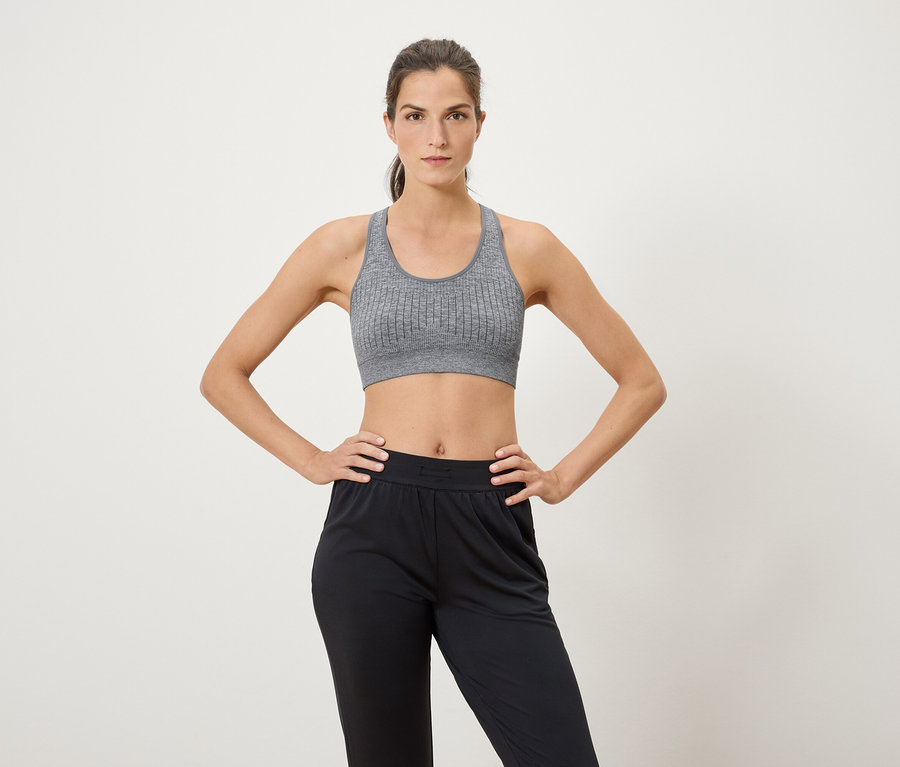 Žena pózuje na bielom pozadí v športovej podprsenke a čiernych nohaviciach.