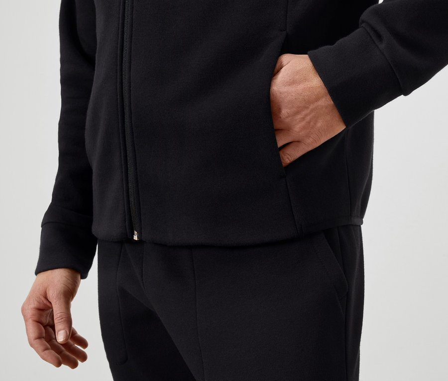 Muž má ruku vo vrecku čiernej bundy na zips a má na sebe čierne nohavice.