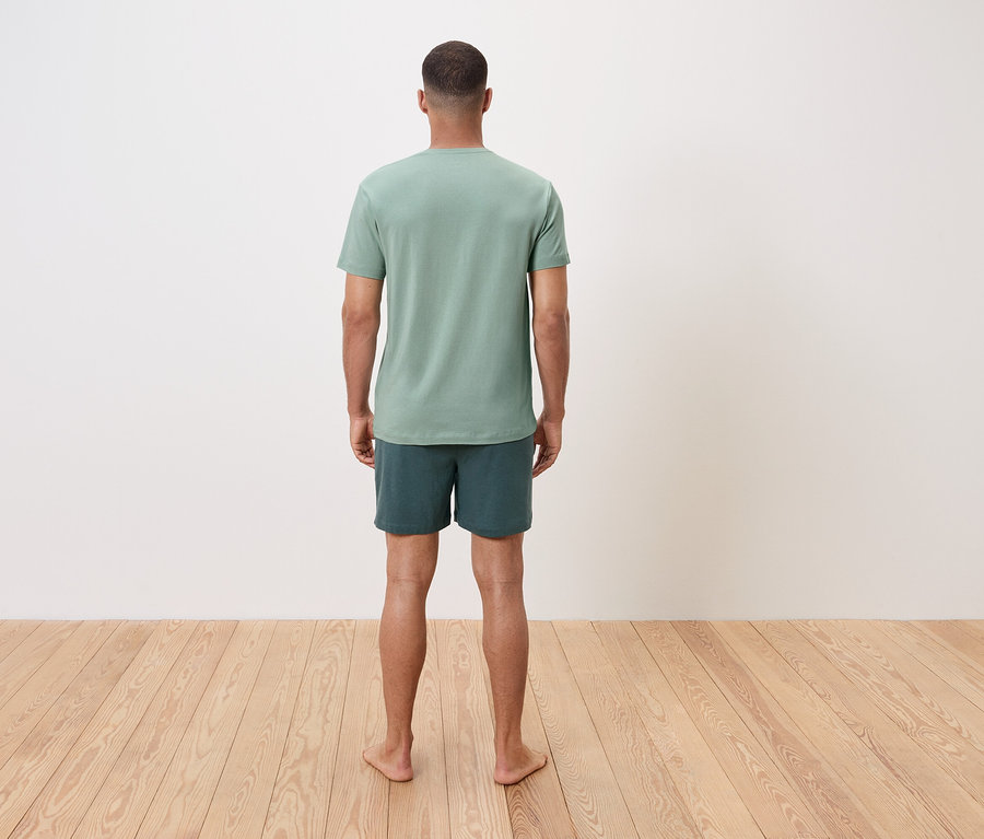 Muž stojí bosý chrbtom k divákovi, má na sebe zelené tričko a šortky.