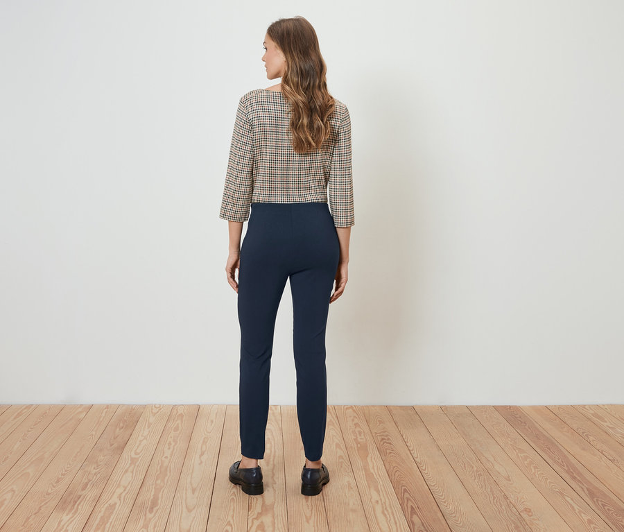 Žena stojí chrbtom k fotoaparátu a má na sebe kockovanú blúzku a tmavomodré nohavice.