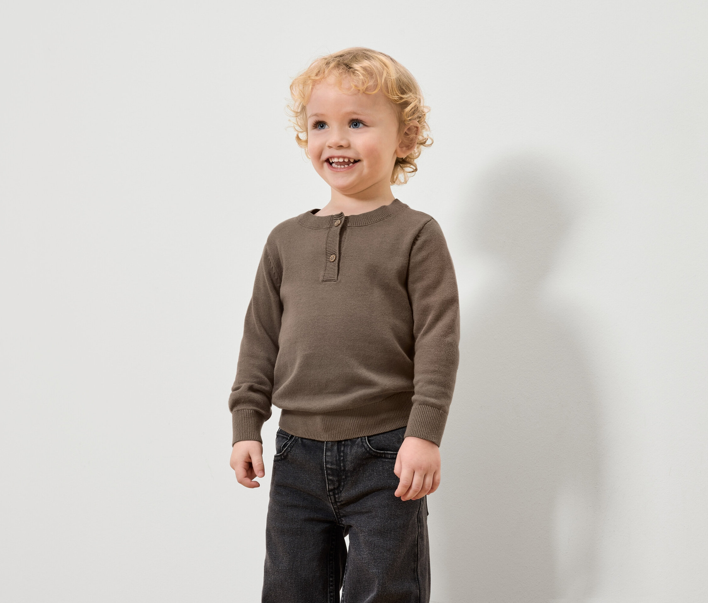 Chlapec s blond kučeravými vlasmi má na sebe hnedý sveter a čierne džínsy.