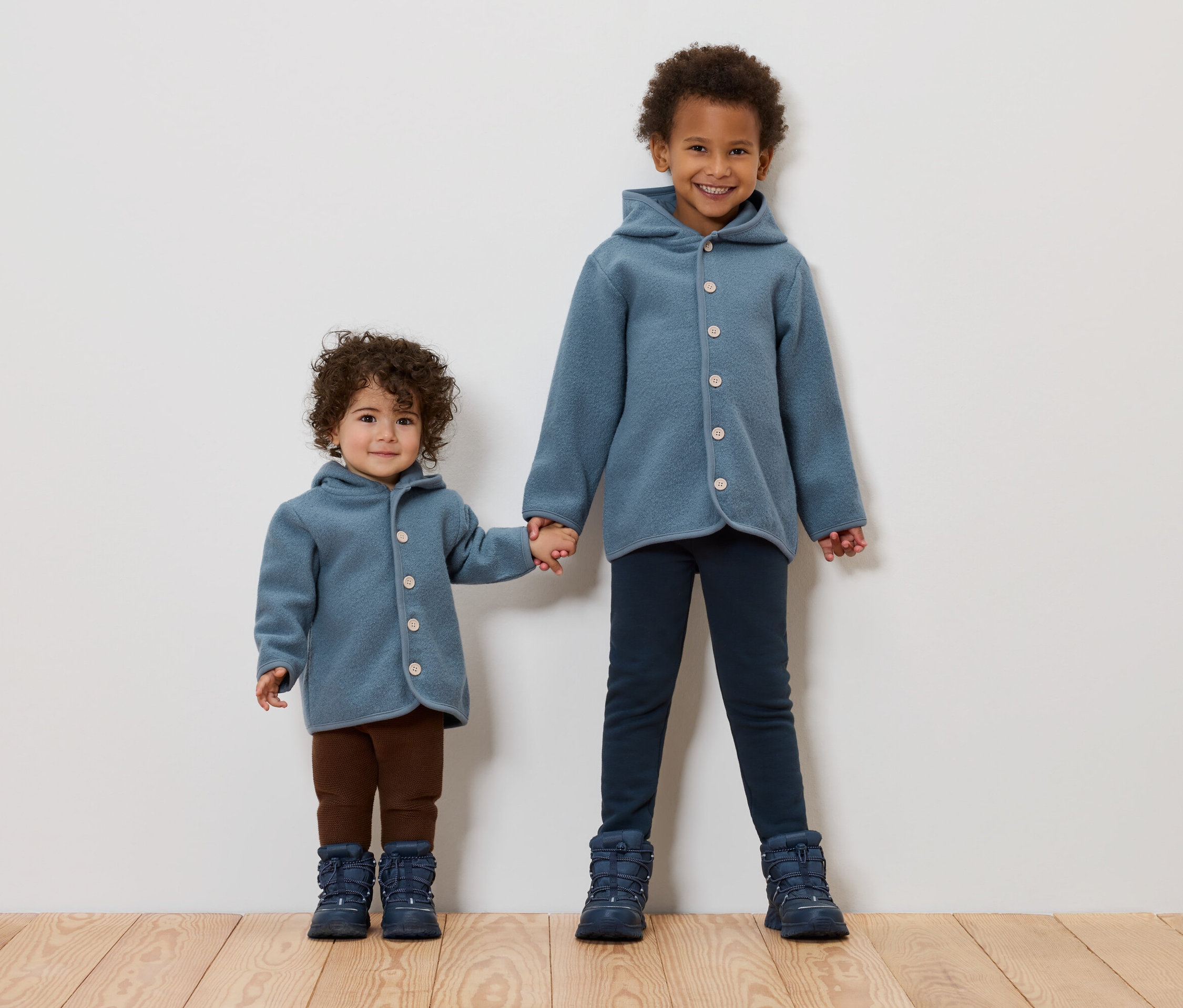 Dve deti sa držia za ruky. Obe majú na sebe modré kabáty a topánky.