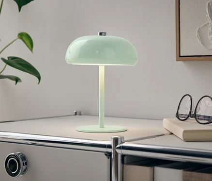 Zelená lampa stojí na bielom nočnom stolíku vedľa rastliny a okuliarov.
