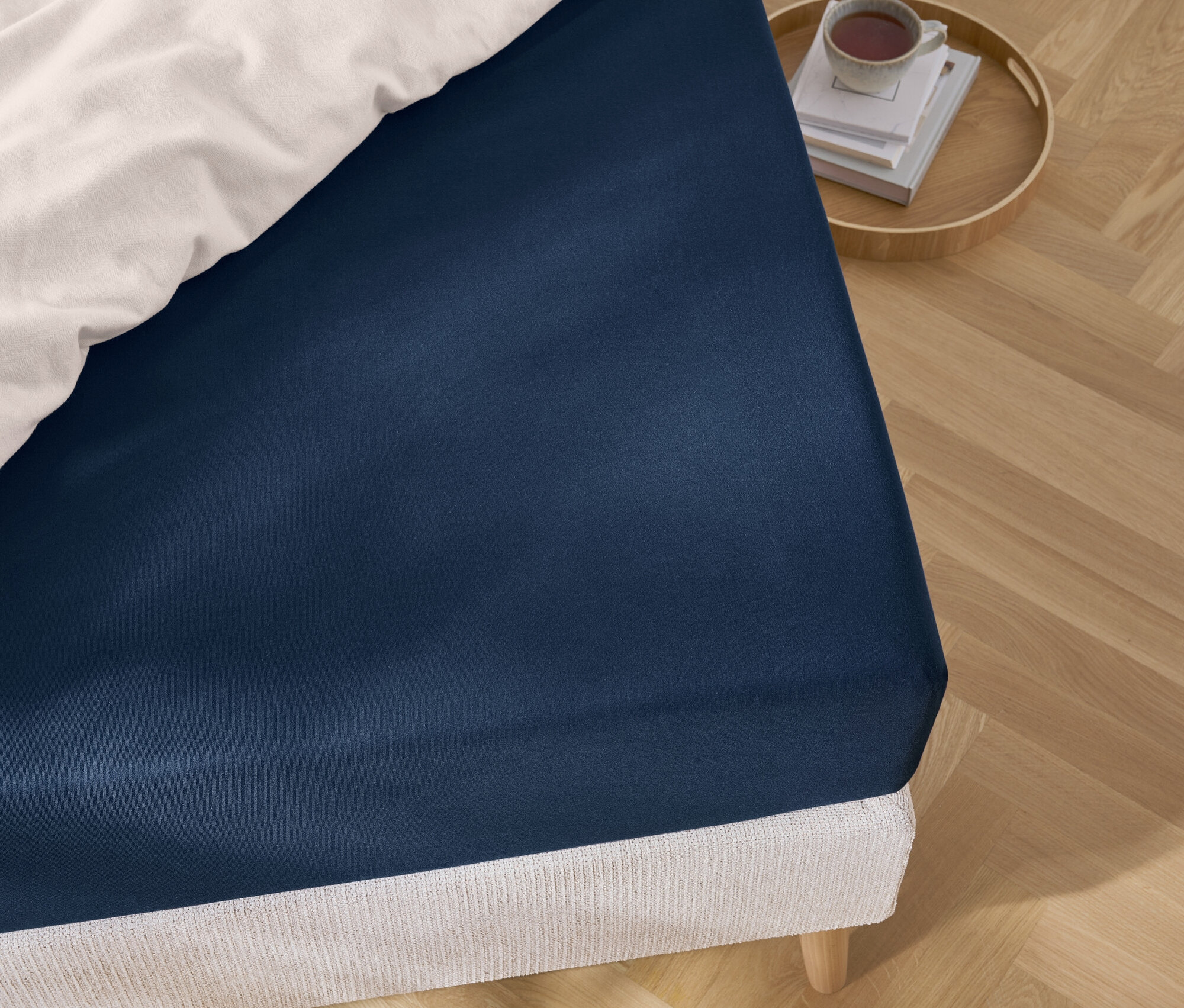 Detail modrej plachty na posteli s bielou posteľnou bielizňou a podnosom s knihami a šálkou.