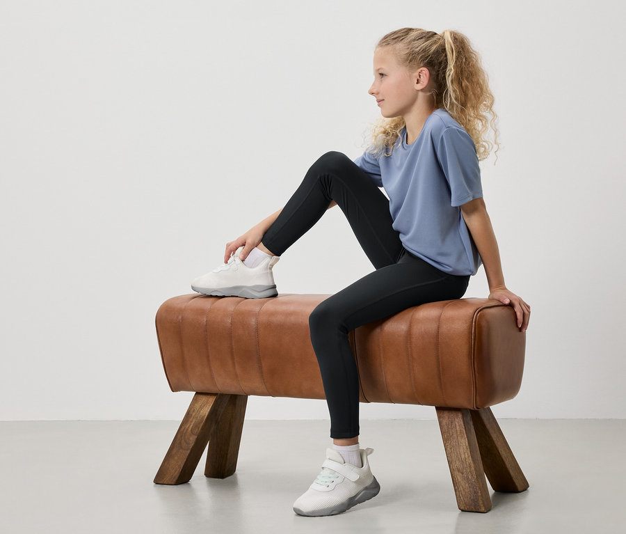 Mladé dievča s blond vlasmi zviazanými do copu sedí na gymnastickom koni. Má na sebe modré tričko, čierne legíny a biele tenisky.