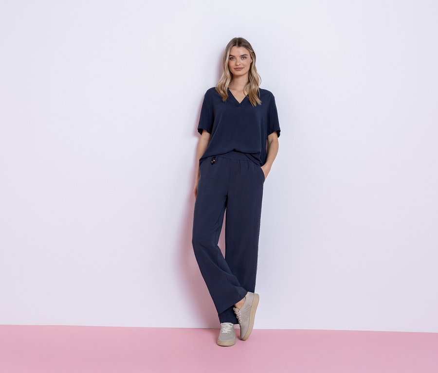 Žena stojí na ružovom a bielom pozadí a má na sebe tmavomodrý outfit pozostávajúci z trička a nohavíc a sivé tenisky.