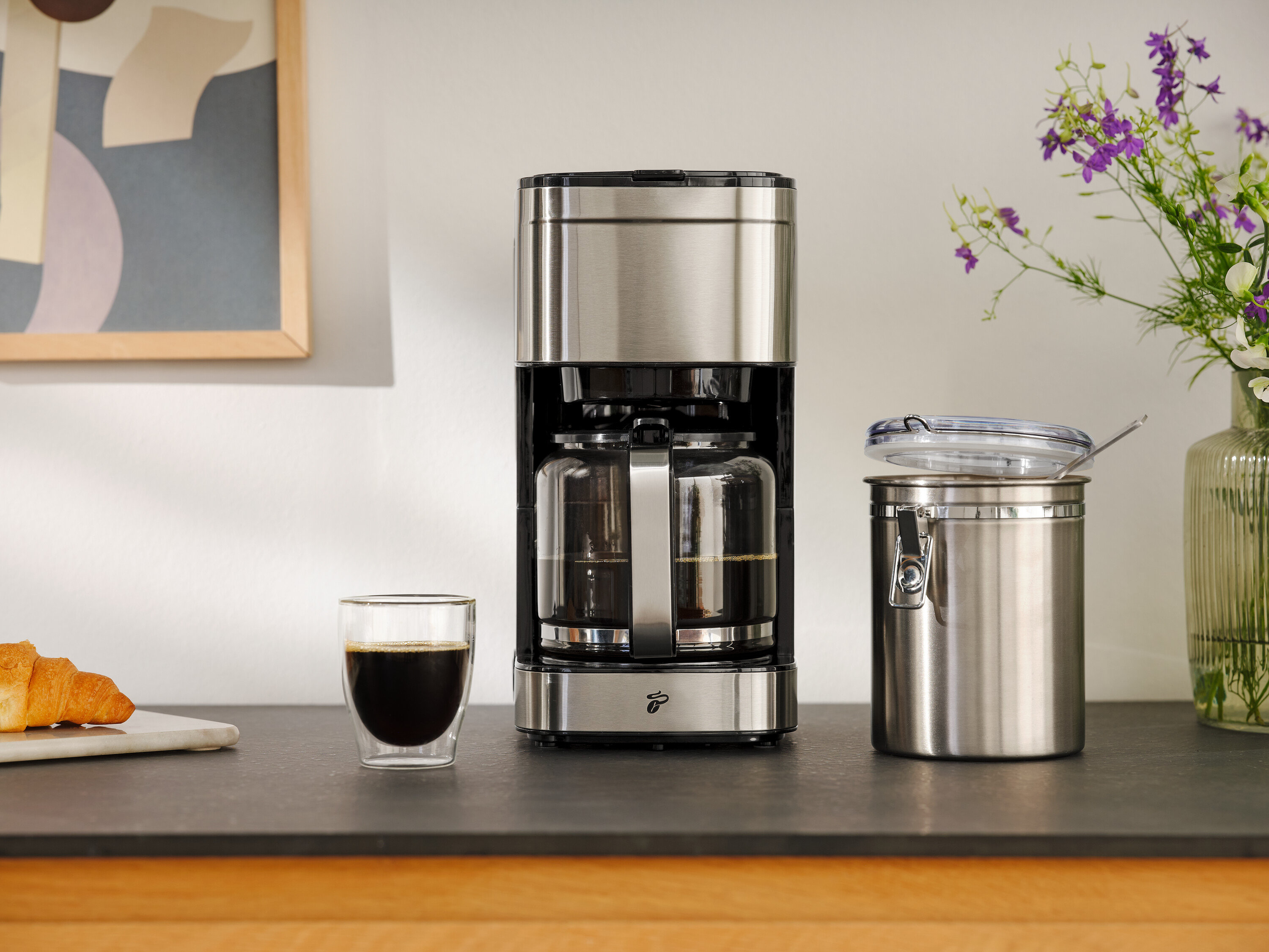 Kávovar vedľa pohára kávy, croissantu a nádoby na uskladnenie.