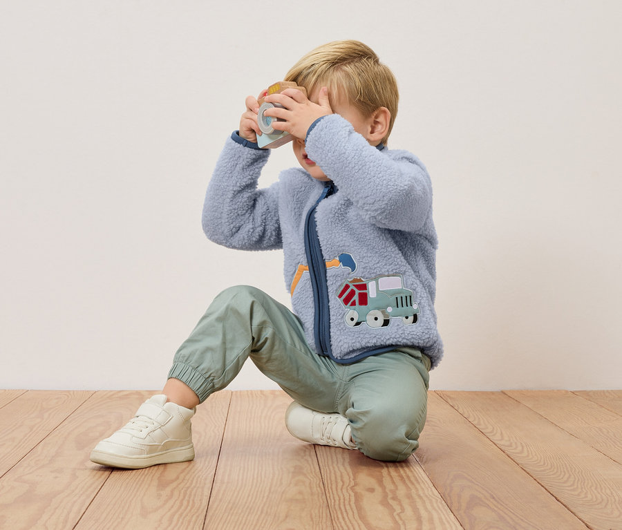 Blond chlapec kľačí na drevenej podlahe a drží pred tvárou hračkársky fotoaparát. Má na sebe modrú fleecovú bundu s aplikáciou nákladného auta a zelené nohavice.