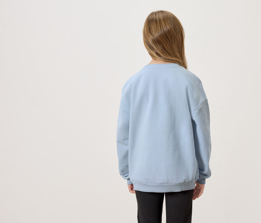 Pohľad zozadu na dieťa s dlhými hnedými vlasmi, oblečené v svetlomodrom svetri a čiernych nohaviciach.