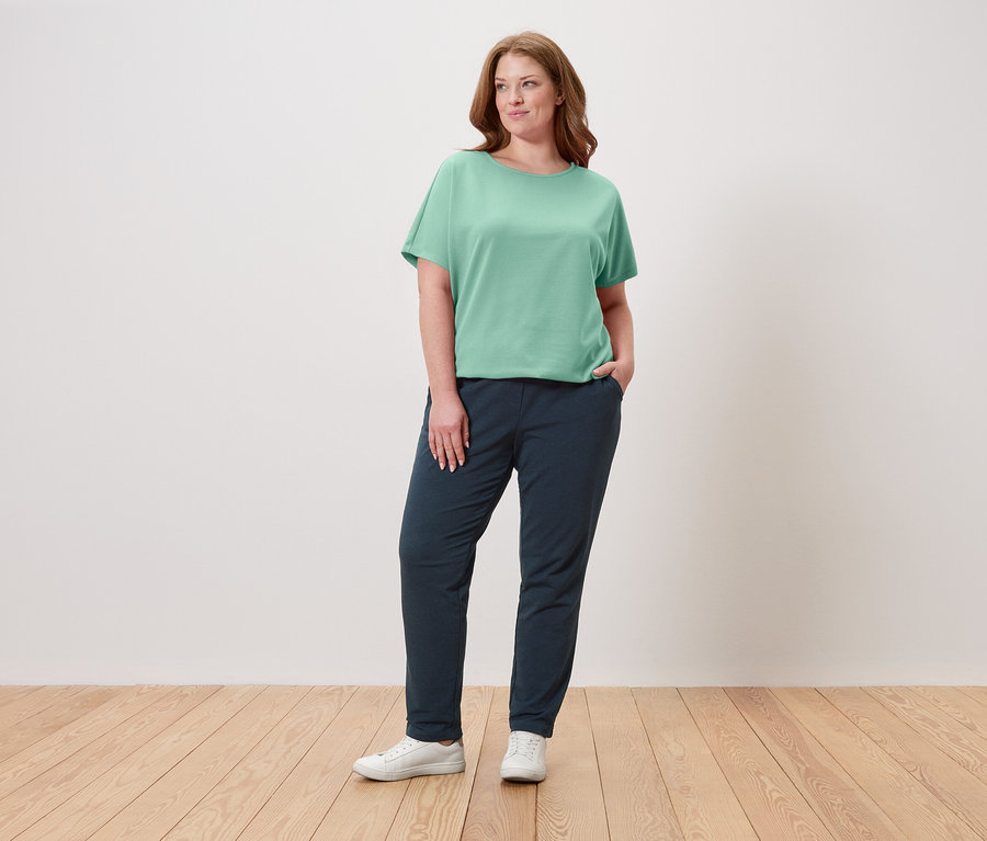 Žena stojí s rukou vo vrecku, má na sebe zelené tričko, tmavomodré nohavice a biele tenisky.