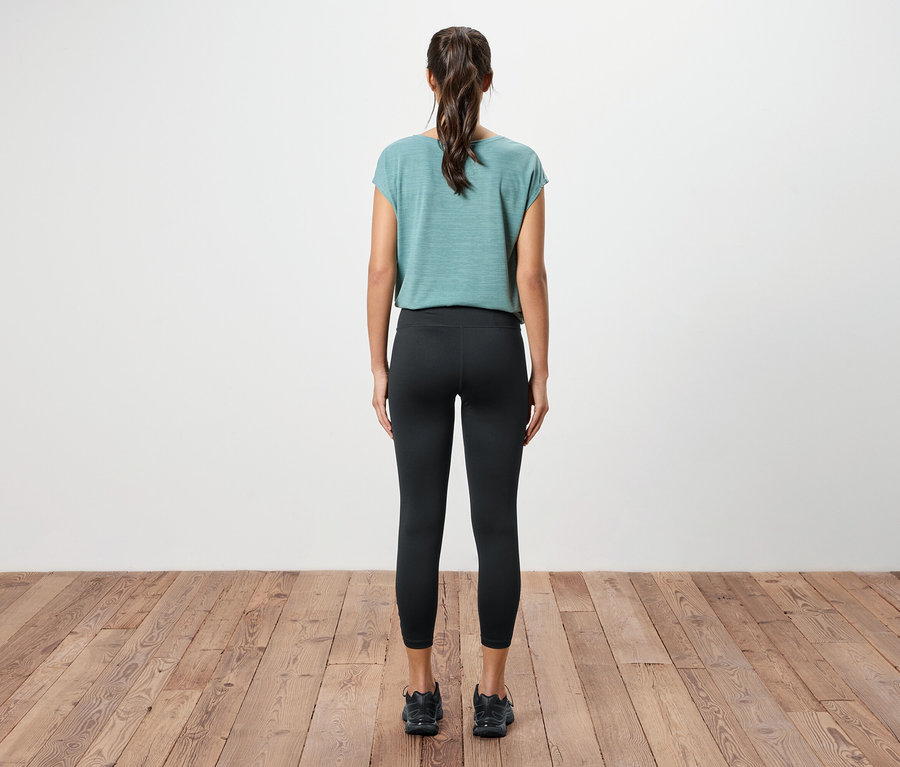 Pohľad zozadu na ženu s konským chvostom, oblečenú v modrozelenom tope, čiernych legínach a čiernych teniskách na drevenej podlahe.