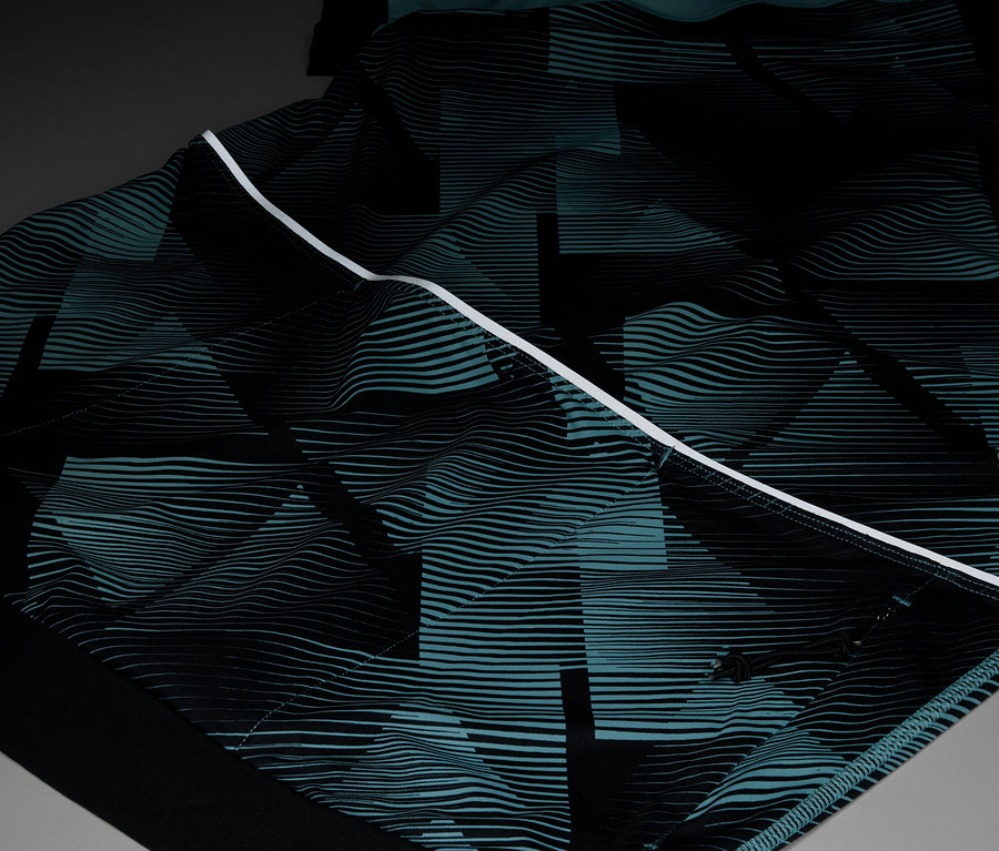 Detail odevu s opakujúcim sa geometrickým vzorom v čiernej a tyrkysovej farbe, zvýraznený bielym pruhom.