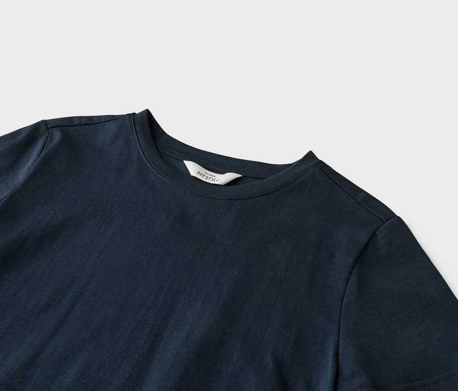 Detailný záber na tmavomodré tričko s bielym štítkom s nápisom "ICHIBO MY STYLE" na golieri, ležiace na svetlosivom pozadí.
