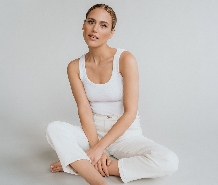 Žena sedí bosá so skríženými nohami na zemi, oblečená v bielom tielku a bielych nohaviciach Angels Denim »Evy Cool« O-Shape.
