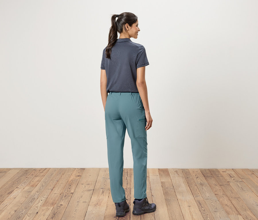 Pohľad zozadu na ženu s konským chvostom, oblečenú v tmavomodrej polokošeli, zelených nohaviciach a turistických topánkach, stojacu pred bielou stenou a drevenou podlahou.