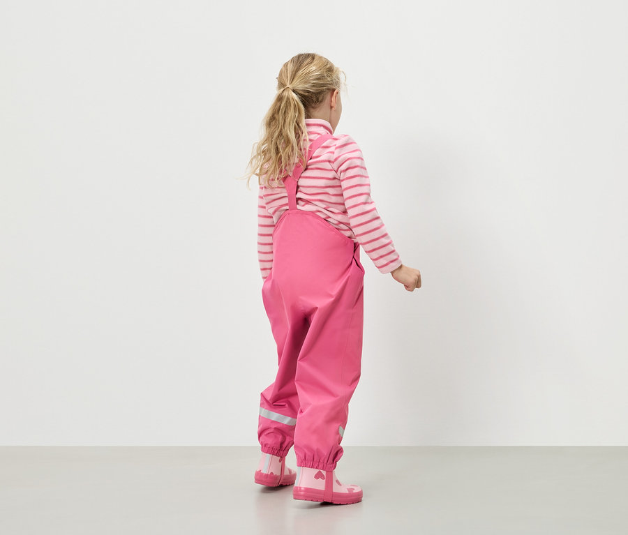 Pohľad zozadu na dievča s blond vlasmi, ktoré má na sebe ružovo-biely pásikavý sveter, ružové dažďové nohavice s trakmi a ružové gumáky so srdiečkami.
