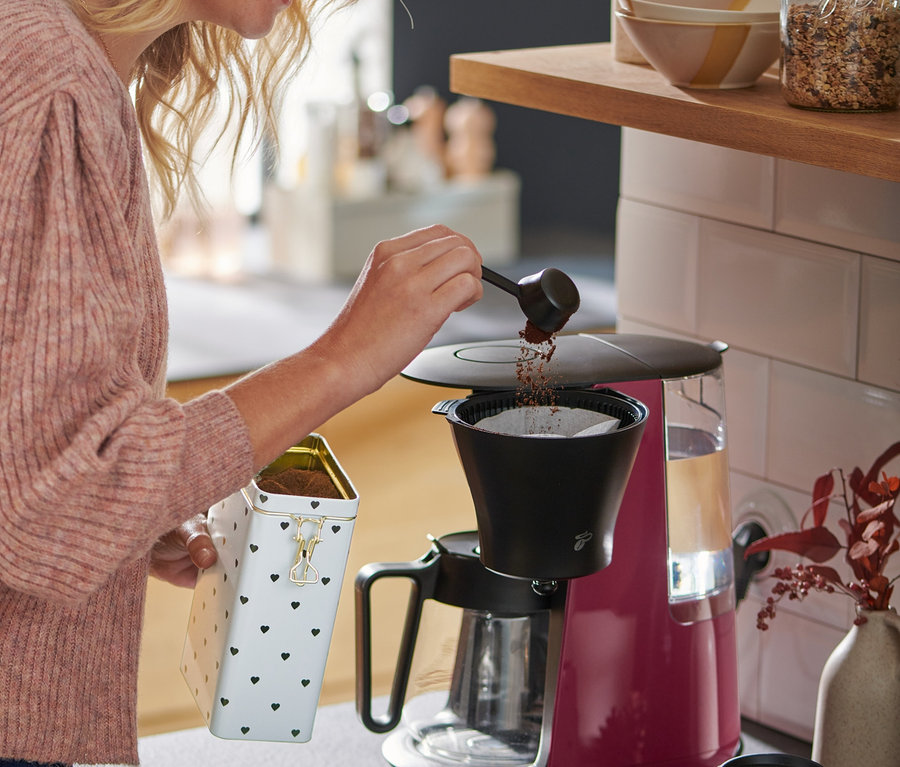Žena s blond vlasmi sype kávu do malinového kávovaru na prekvapkávanú kávu »Let's Brew«.
