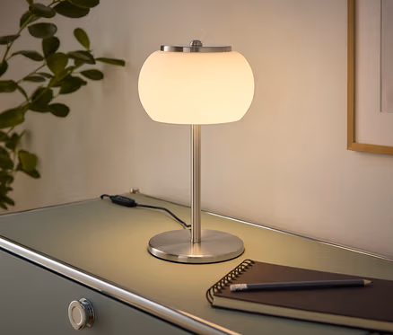 Svietiaca stolová lampa s bielym guľovým tienidlom a brúseným kovovým podstavcom stojí na svetlozelenej komode. Vedľa nej leží zápisník a ceruzka. Vľavo je rastlina, vpravo zarámovaný obraz na stene.